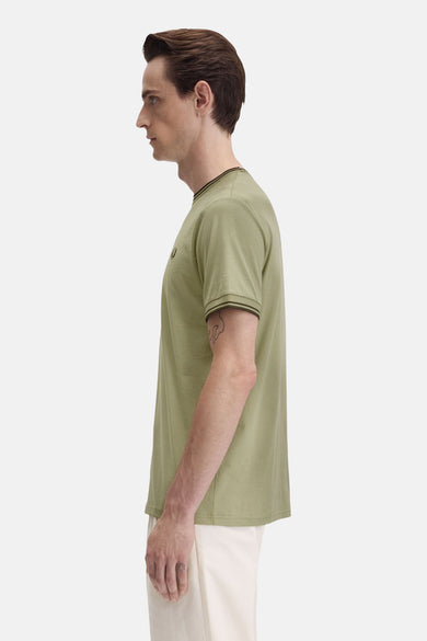 T-shirt olive à manches courtes de Fred Perry avec logo brodé, porté avec un pantalon blanc.