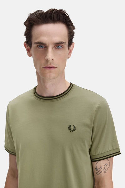 T-shirt olive à manches courtes de FRED PERRY, avec col et bordures rayés, logo brodé et tatouage discret sur le bras.
