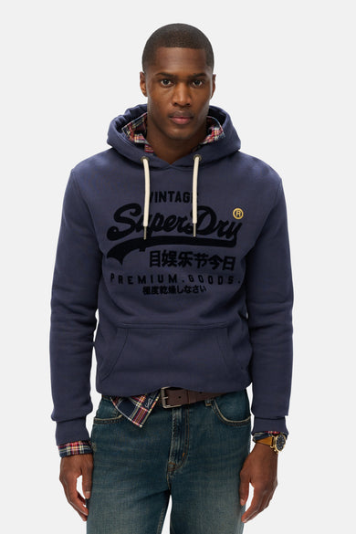 Sweat à capuche bleu de Superdry, avec motif imprimé, porté sur une chemise à carreaux et un jean.
