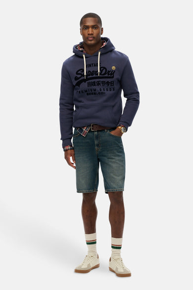 Sweat à capuche bleu par SUPERDRY, avec doublure à carreaux et motif imprimé, porté avec des shorts en jean et une chemise à carreaux.