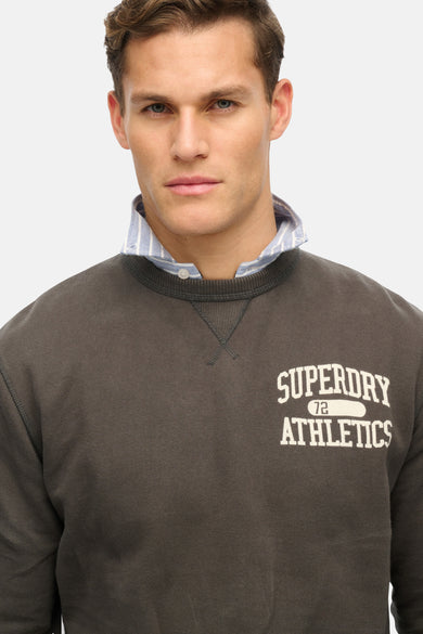 Sweat col rond gris de SUPERDRY, porté avec une chemise à rayures bleues et blanches.