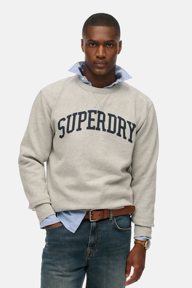 Sweat col rond gris de SUPERDRY, porté sur une chemise bleue, avec un jean et une ceinture marron.