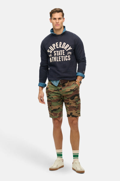 Sweat col rond bleu de SUPERDRY, porté avec une chemise en jean et un short camouflage.