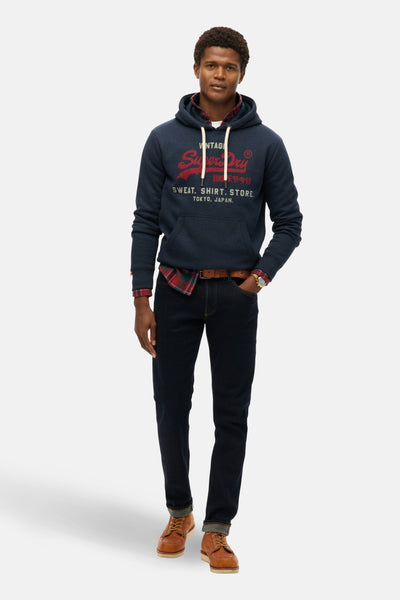 Sweat à capuche bleu marine de Superdry, porté sur une chemise à carreaux avec un jean et des chaussures marron.
