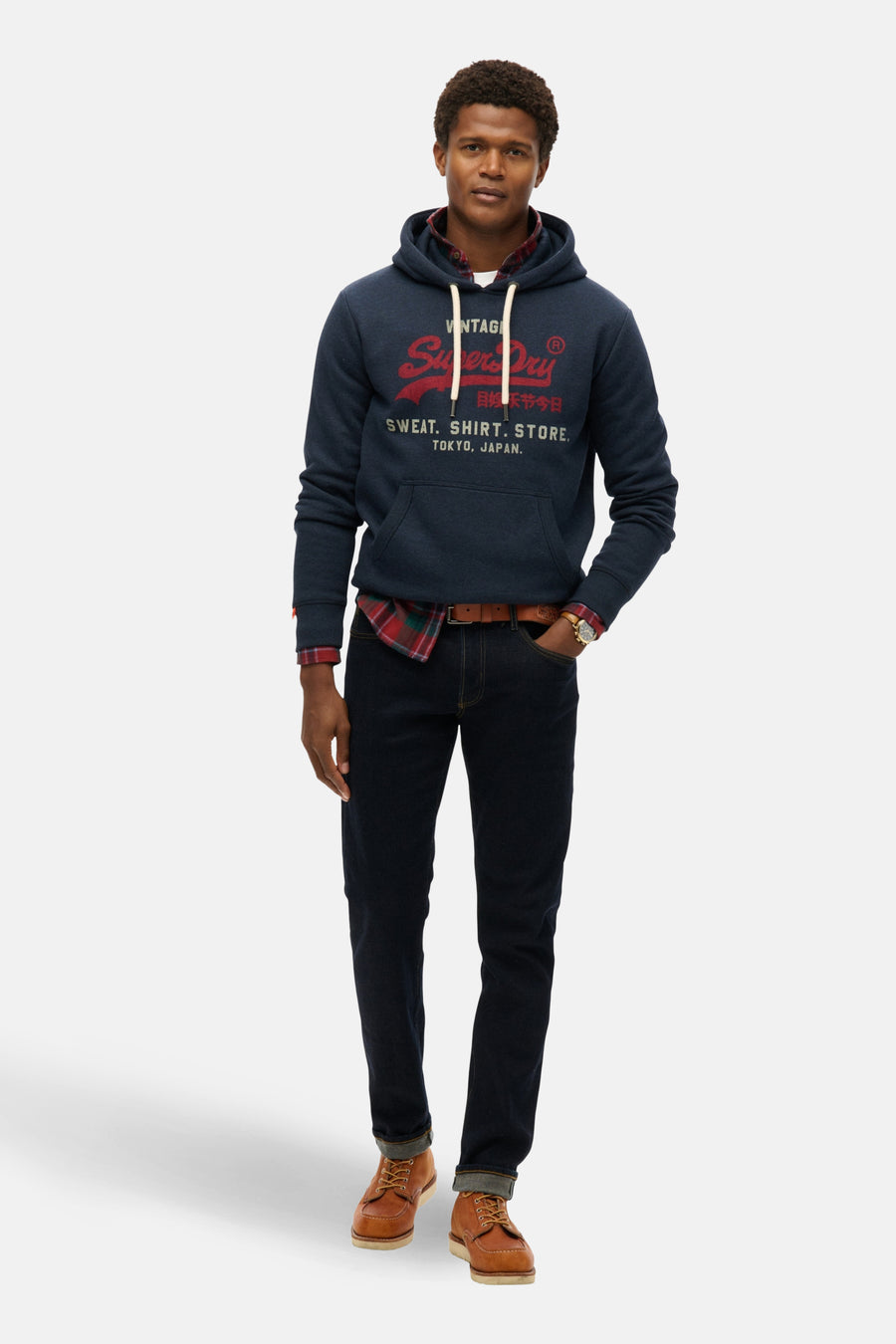 Sweat à capuche bleu marine de Superdry, porté sur une chemise à carreaux avec un jean et des chaussures marron.
