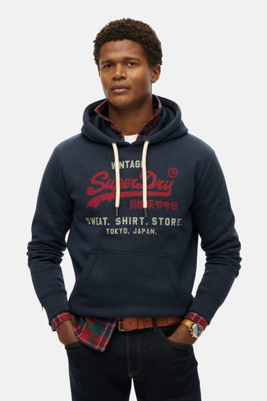 Sweat à capuche bleu chiné de SUPERDRY, porté sur une chemise à carreaux, avec jean et montre.
