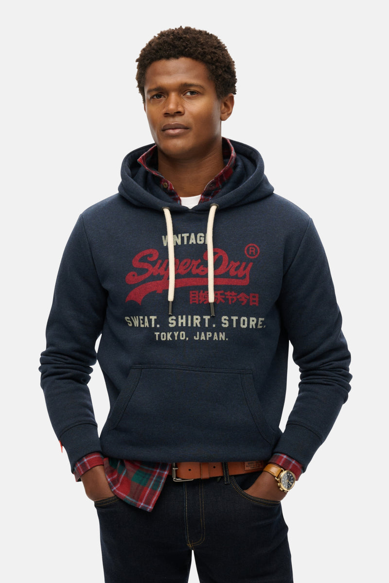 Sweat à capuche bleu chiné de SUPERDRY, porté sur une chemise à carreaux, avec jean et montre.
