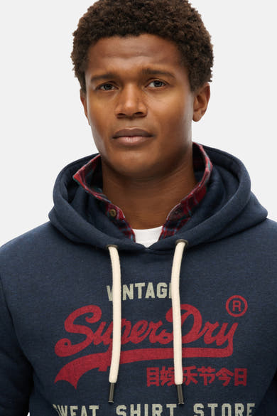 Sweat à capuche bleu chiné de Superdry, porté sur une chemise à carreaux rouge et marine.