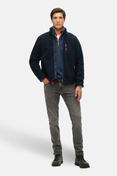 Blauwe bodywarmer van SUPERDRY, gedragen over een geruit overhemd met grijze jeans en veterschoenen.