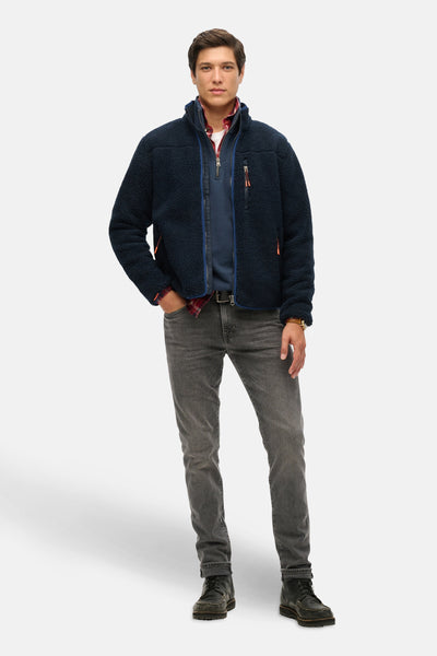 Blauwe bodywarmer van SUPERDRY, gedragen over een geruit overhemd met grijze jeans en veterschoenen.