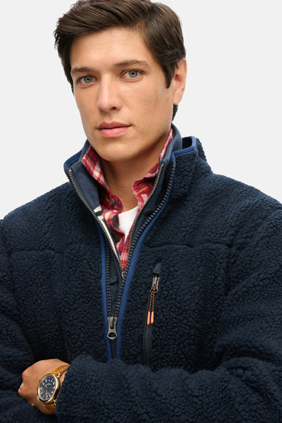 Blauwe bodywarmer van zachte stof van SUPERDRY, gedragen over een geruit overhemd en een wit T-shirt.