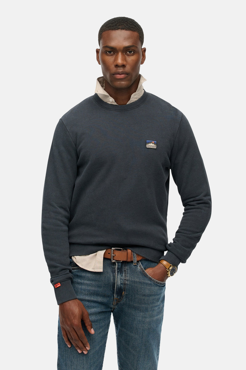 Marineblauwe trui van SUPERDRY, gedragen met een gebroken witte blouse, een spijkerbroek en een bruine riem.