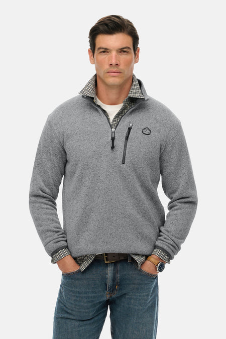Grijze fleece van SUPERDRY, gedragen over een geruit overhemd, met ritssluiting tot halverwege en een blauwe spijkerbroek.
