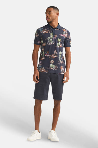 Multikleurig poloshirt van Petrol Industries met woestijnpatroon en cactussen, gedragen door een man met een donkere korte broek en witte sneakers.