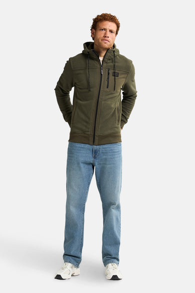 Groen vest met rits van Petrol Industries, met capuchon en ritszakken, gedragen met een blauwe jeans en witte sneakers.