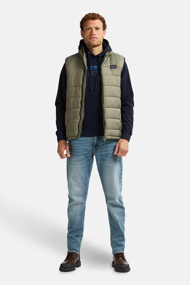 Kaki mouwloos vest van Petrol Industries, gedragen met een marineblauwe hoodie en een spijkerbroek.