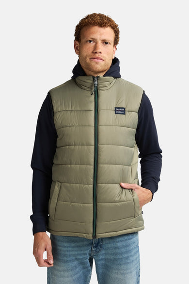 Kaki mouwloos vest van Petrol Industries, gedragen over een marineblauwe hoodie en een blauwe jeans.