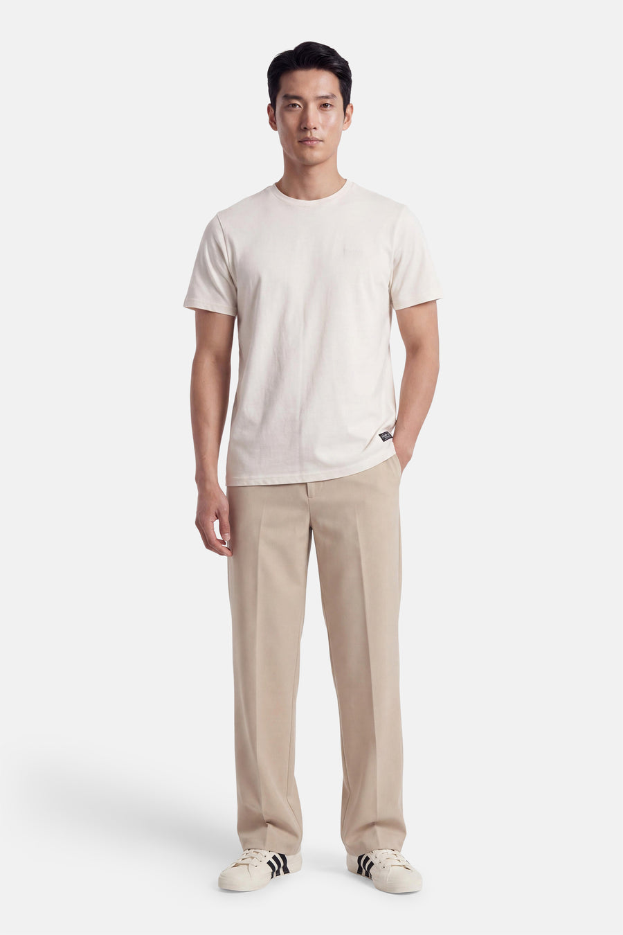 Ecru short-sleeve t-shirt with round neckline and small chest logo, worn by model in full-length front view with beige trousers and white sneakers.
