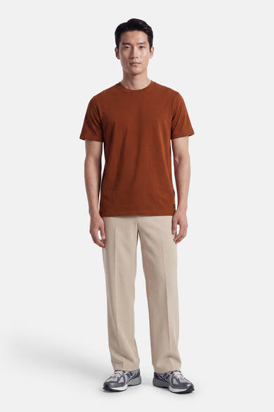 Orange short-sleeve t-shirt with round neckline and small chest logo, worn by model in full-length front view with beige trousers and sneakers.