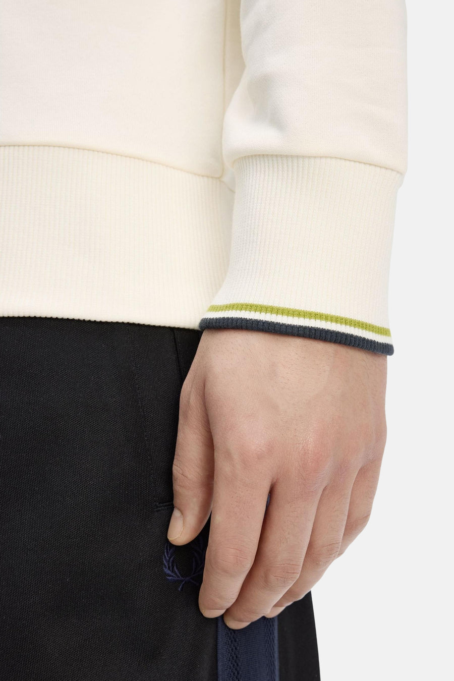 Sweat col montant écru de Fred Perry, détail rayé aux poignets, porté avec un pantalon noir à logo discret.
