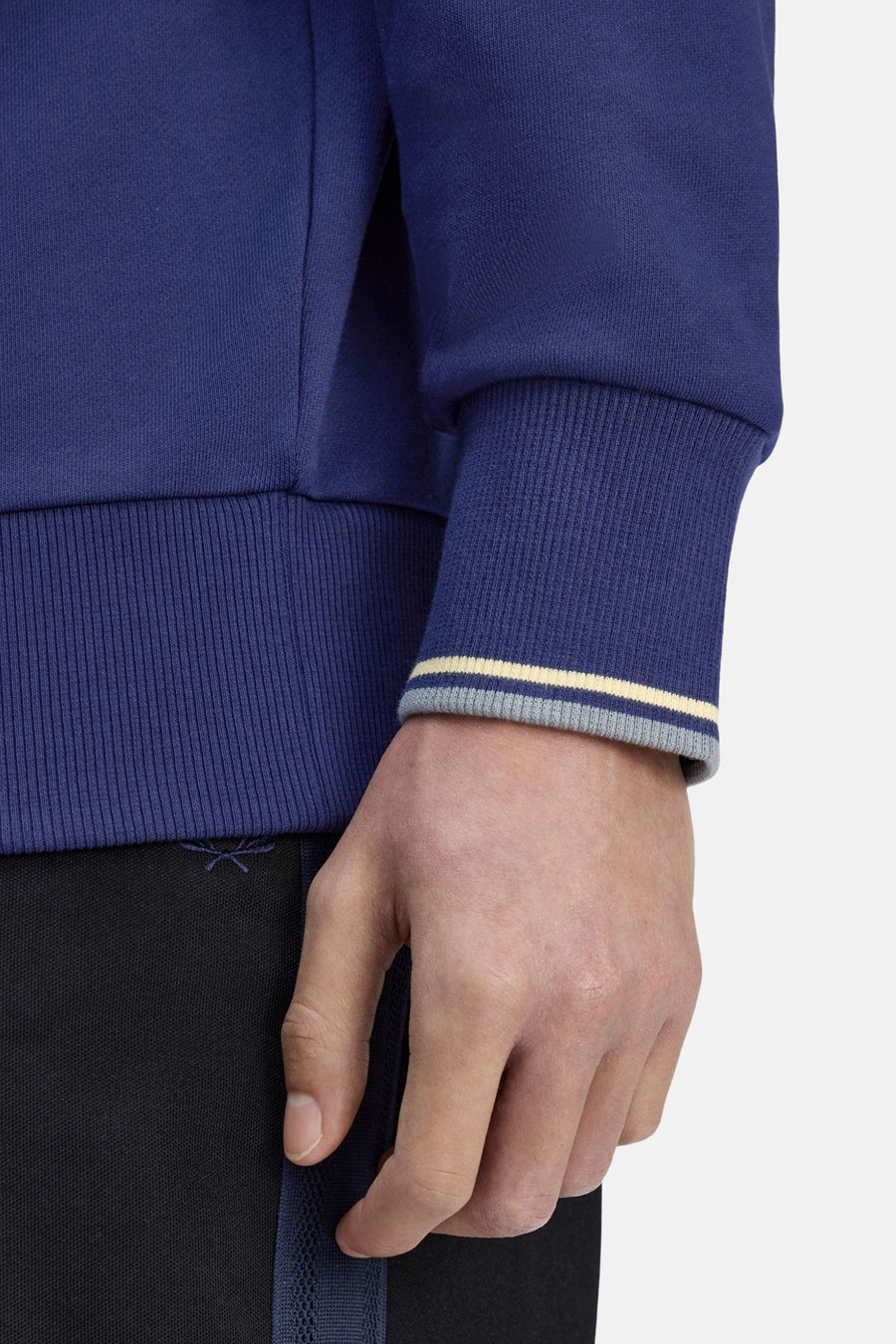 Sweat col roulé bleu de FRED PERRY avec poignet côtelé à rayures et pantalon noir visible.