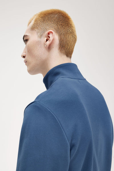 Blauwe Fred Perry sweater met opstaande kraag, gedragen door een man met kort blond haar en een oorbel.