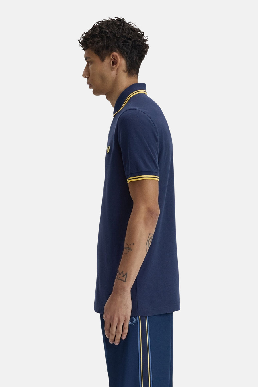 Polo bleu à manches courtes de Fred Perry, avec rayures jaunes au col et aux manches, porté avec un pantalon de survêtement bleu.
