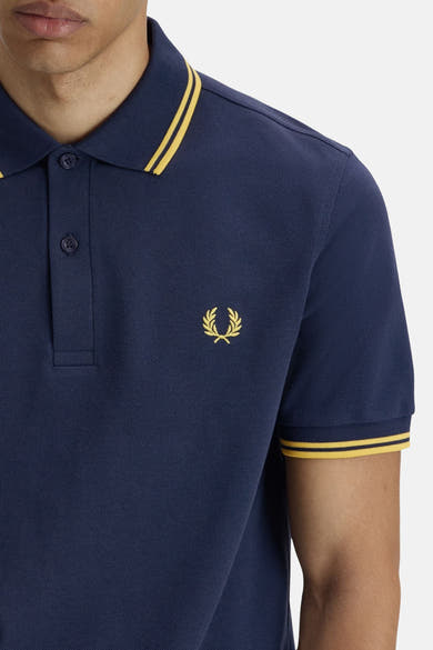 Polo à manches courtes bleu de Fred Perry, avec rayures jaunes au col et aux manches, et logo brodé.