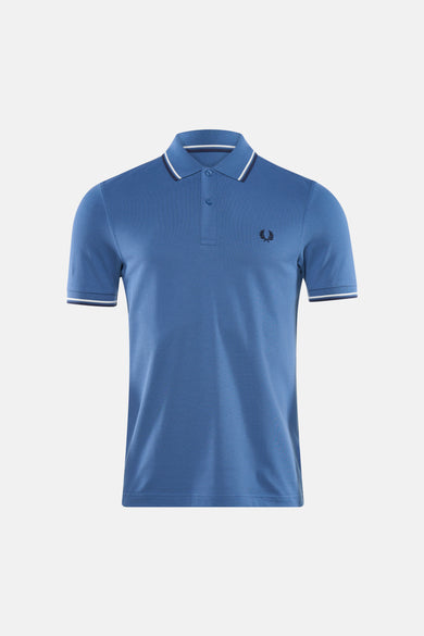 Blauwe FRED PERRY polo, met witte gestreepte details op de kraag en mouwen, gedragen met een lichte broek en loafers.