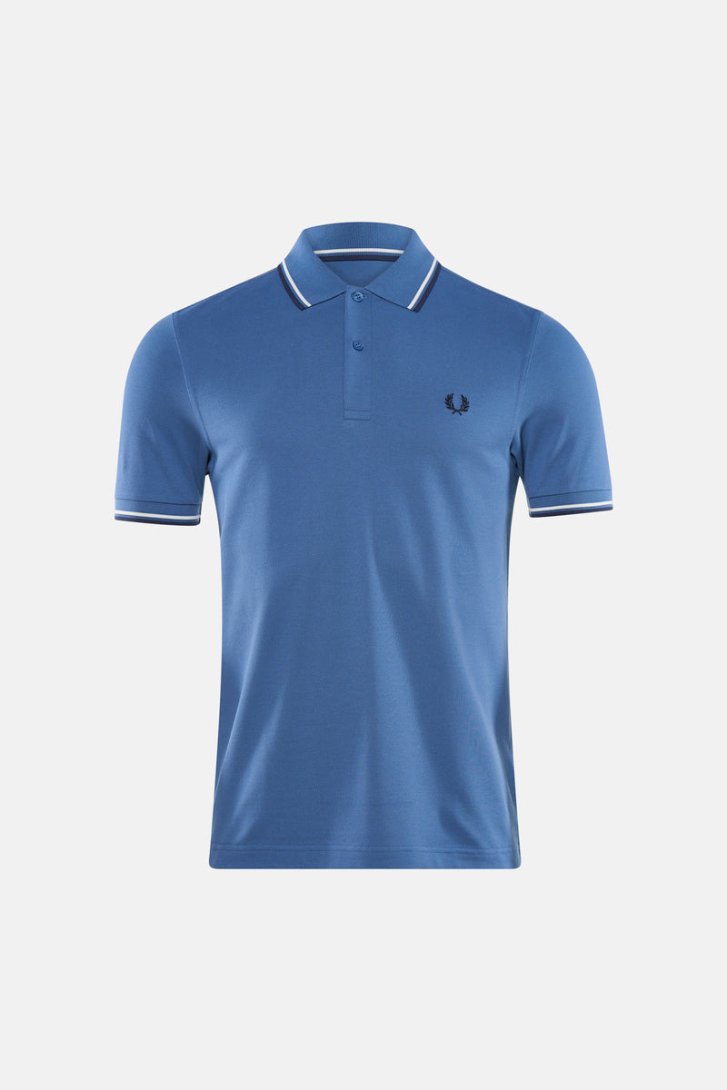Blauwe FRED PERRY polo, met witte gestreepte details op de kraag en mouwen, gedragen met een lichte broek en loafers.