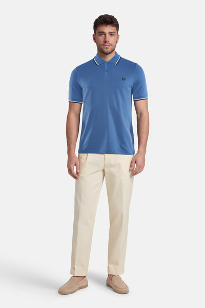 Polo bleu FRED PERRY, avec des détails rayés blancs au col et aux manches, porté avec un pantalon clair et des mocassins.
