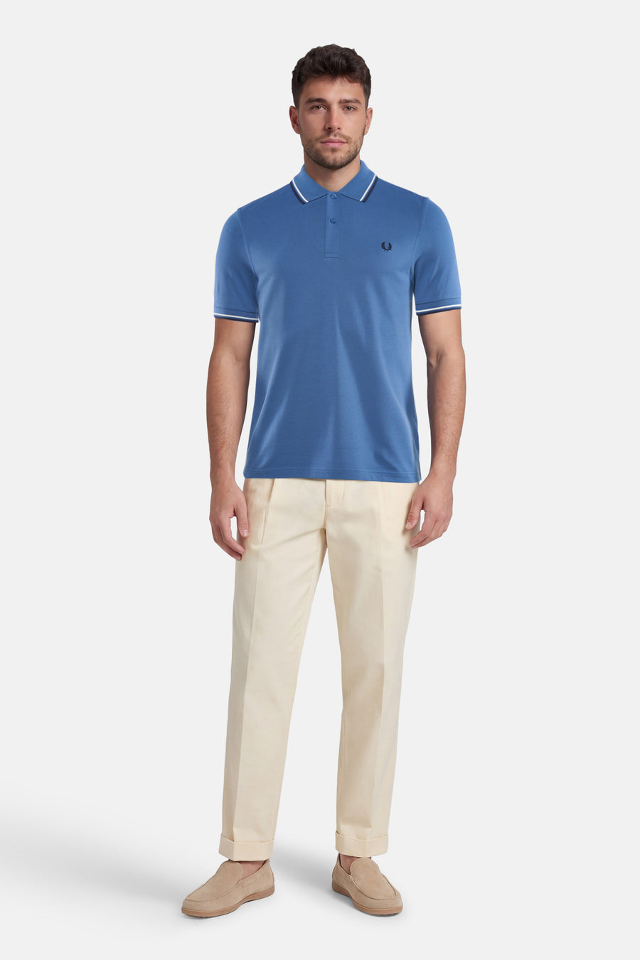 Polo bleu FRED PERRY, avec des détails rayés blancs au col et aux manches, porté avec un pantalon clair et des mocassins.

