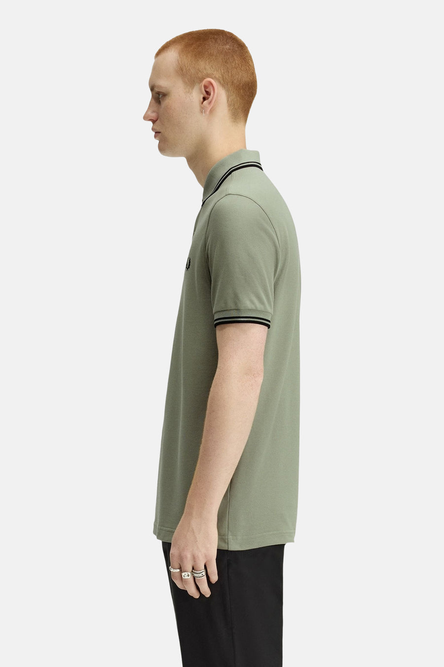 Polo à manches courtes vert de FRED PERRY, col et manches à rayures noires, porté avec un pantalon noir et des bagues.