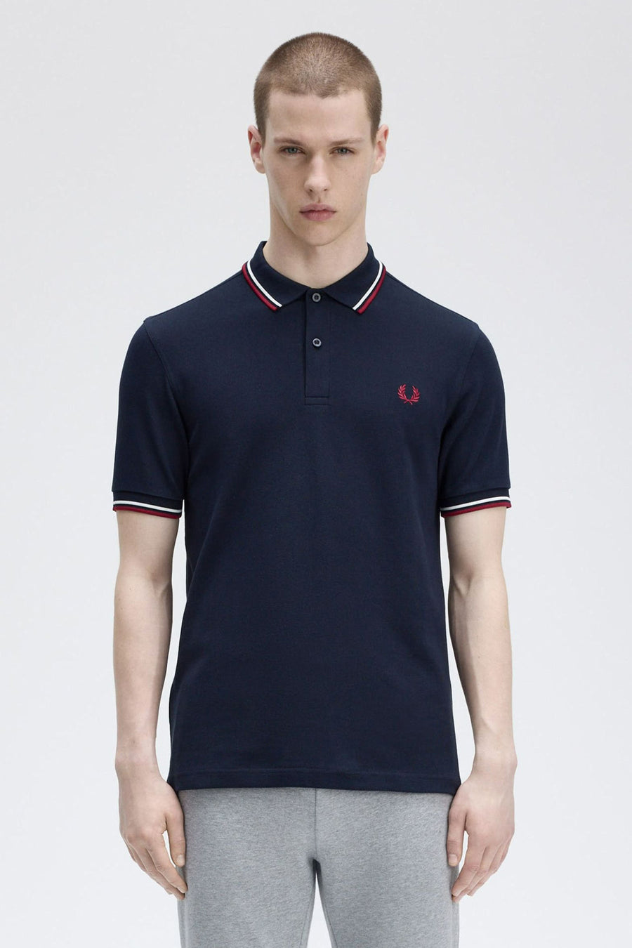 Polo bleu à manches courtes de Fred Perry, avec col et manches rayés en blanc et rouge, porté avec un pantalon gris.
