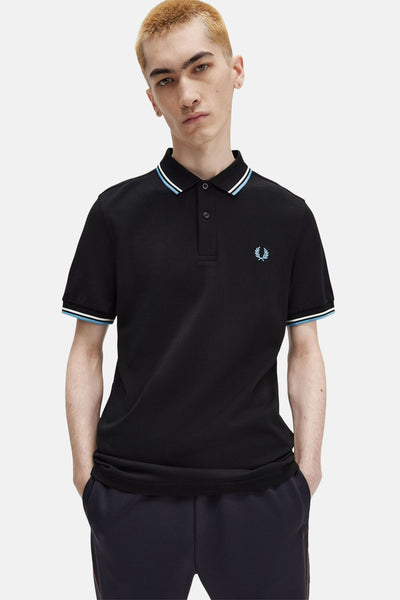 Polo noir à manches courtes de Fred Perry, avec rayures contrastées au col et aux manches, porté avec un pantalon marine.

