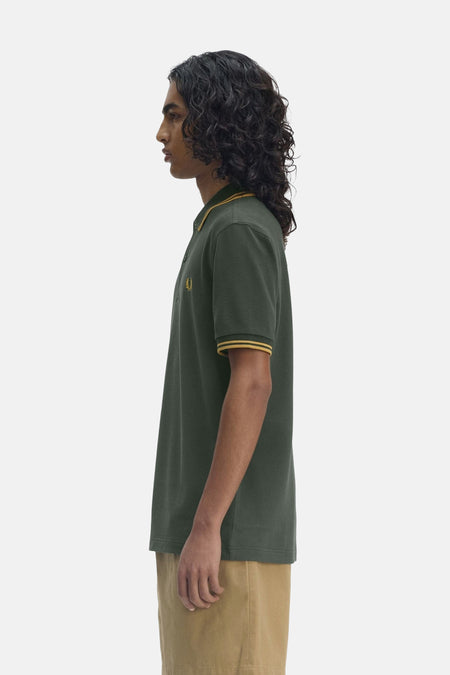 Polo vert à manches courtes de FRED PERRY, avec bordures jaune doré au col et aux manches, porté avec un pantalon beige.