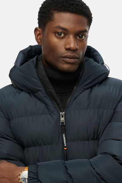 Veste courte bleue de SUPERDRY, avec capuche et fermeture éclair apparente, portée sur un col roulé noir.