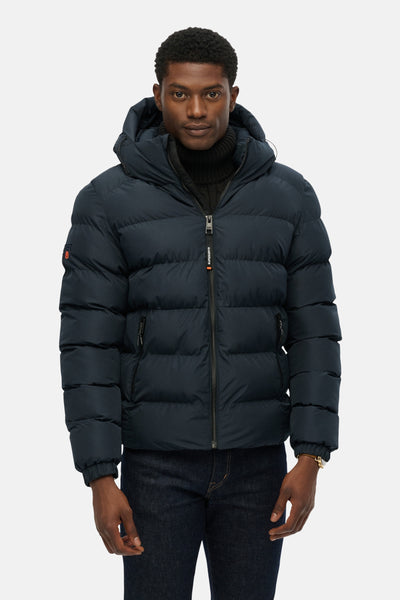 Veste courte bleue de SUPERDRY, modèle matelassé à capuche, portée avec un pull col roulé noir et un jean foncé.
