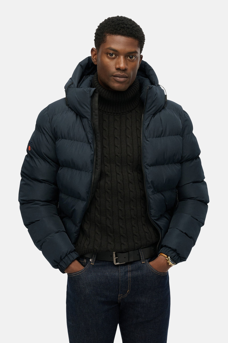 Veste courte bleue de SUPERDRY, matelassée et à capuche, portée sur un pull à col roulé et un jean.