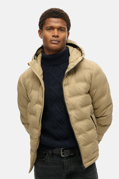 Veste de pluie beige de SUPERDRY, portée par un homme avec un pull en maille torsadée et un jean noir.