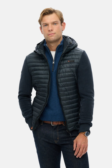 Veste de pluie bleue par SUPERDRY, avec fermeture éclair, capuche et poches, portée sur un pull bleu et un jean.
