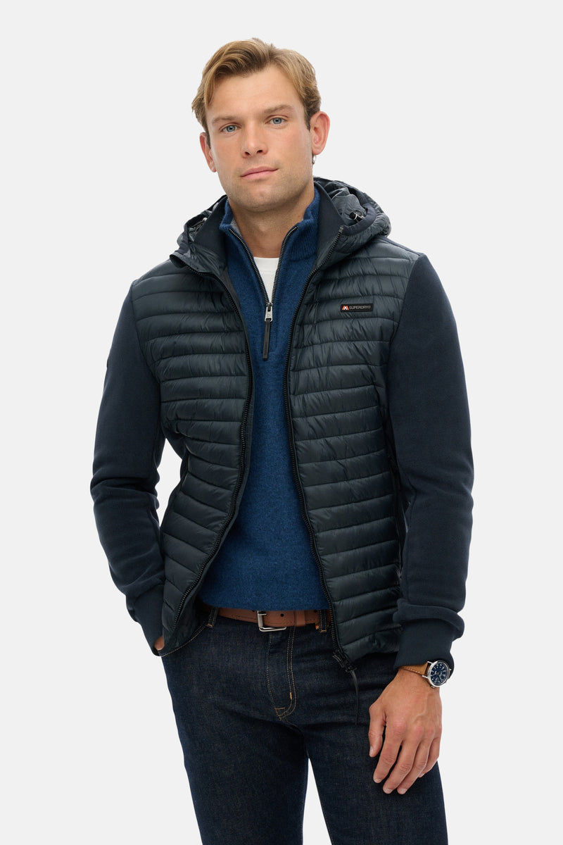 Veste de pluie bleue par SUPERDRY, avec fermeture éclair, capuche et poches, portée sur un pull bleu et un jean.
