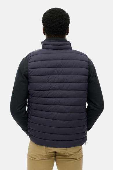 Veste sans manches bleu de SUPERDRY, matelassée avec un col montant, portée sur un pull sombre et un pantalon beige, vue de dos.