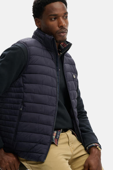 Blauwe mouwloze jas van SUPERDRY, gewatteerd met ritssluiting, gedragen over een zwart poloshirt en een beige broek.
