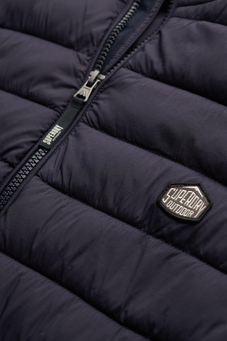 Blauwe mouwloze jas van SUPERDRY, gewatteerd met ritssluitingdetails en een merklogo.