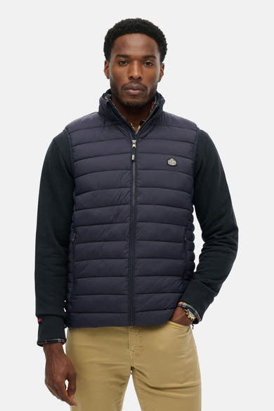 Blauwe mouwloze jas van SUPERDRY, met ritssluiting over een gebreide trui en beige broek.