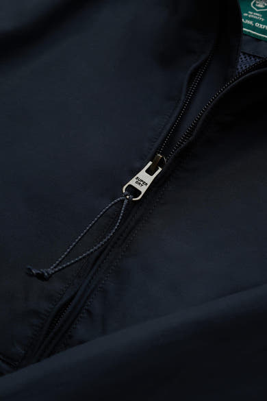 Veste de pluie bleue de SUPERDRY, gros plan sur le tirette métallique avec logo et cordon de serrage.