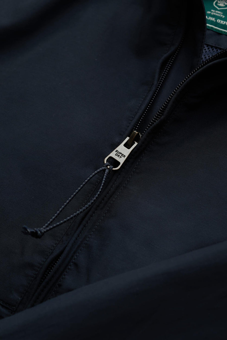 Veste de pluie bleue de SUPERDRY, gros plan sur le tirette métallique avec logo et cordon de serrage.