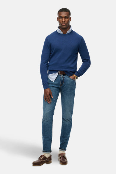 Pull col rond bleu par SUPERDRY, porté sur une chemise rayée, avec jean et chaussures bateau marron.
