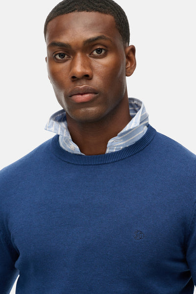 Pull col rond bleu de SUPERDRY, porté avec une chemise rayée à col boutonné.